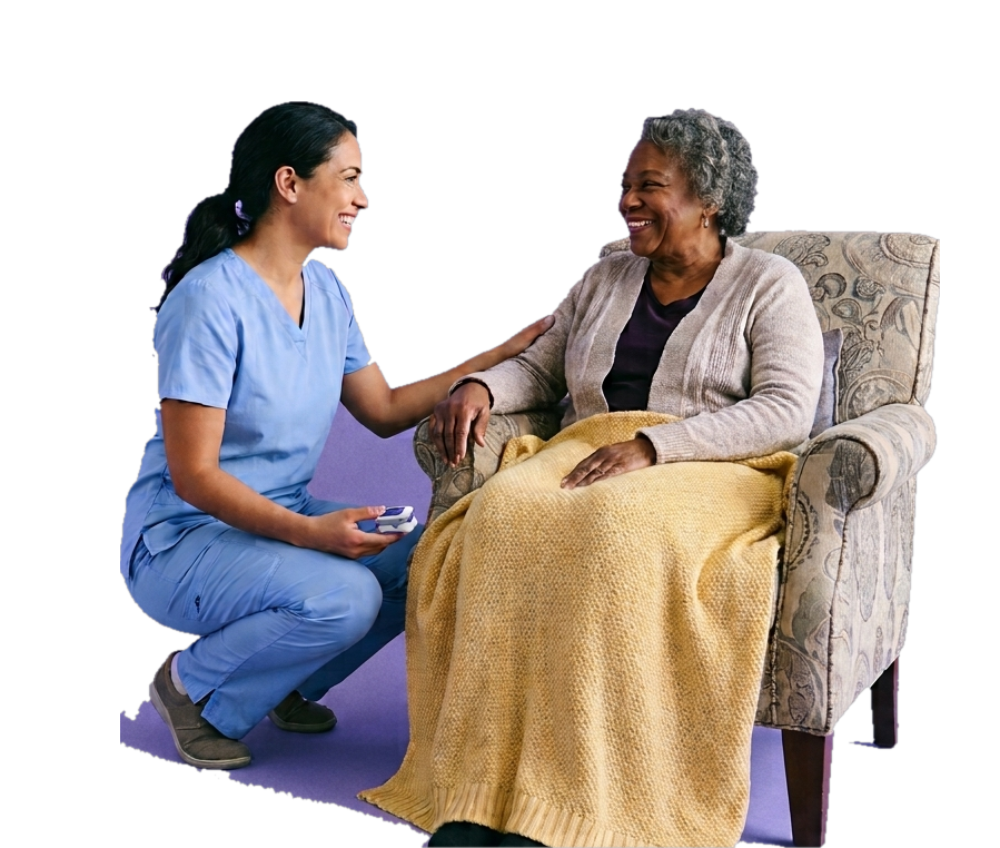 A home health nurse in blue scrubs crouched beside an older adult seated in an armchair with a yellow throw, both smiling.